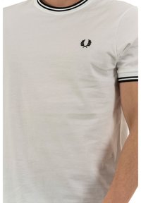 White crew neck t-shirt in soft cotton with black ribbed trim on collar and sleeves. Features a small black embroidered logo on the chest.