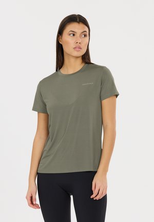 Vrouw met stijl donker haar die een effen olijfgroen sportshirt met korte mouwen en zwarte leggings draagt, kijkt naar links.
