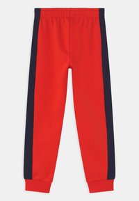 Červené tepláky s navy pruhy na boku, vybavené elastickým pasem a žebrovanými manžetami pro těsné přilnutí. Měkká textura látky.