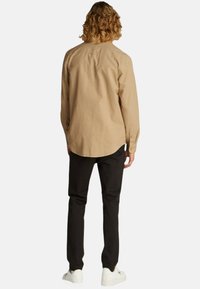 Camicia a maniche lunghe beige con orlo arcuato e polsini a bottone, abbinata a pantaloni neri e sneakers bianche, vista da dietro.