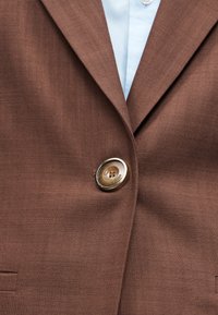 Blazer marrone testurizzato abbottonato su una camicia azzurro chiaro con colletto, mostrando il rever e un singolo bottone marrone.