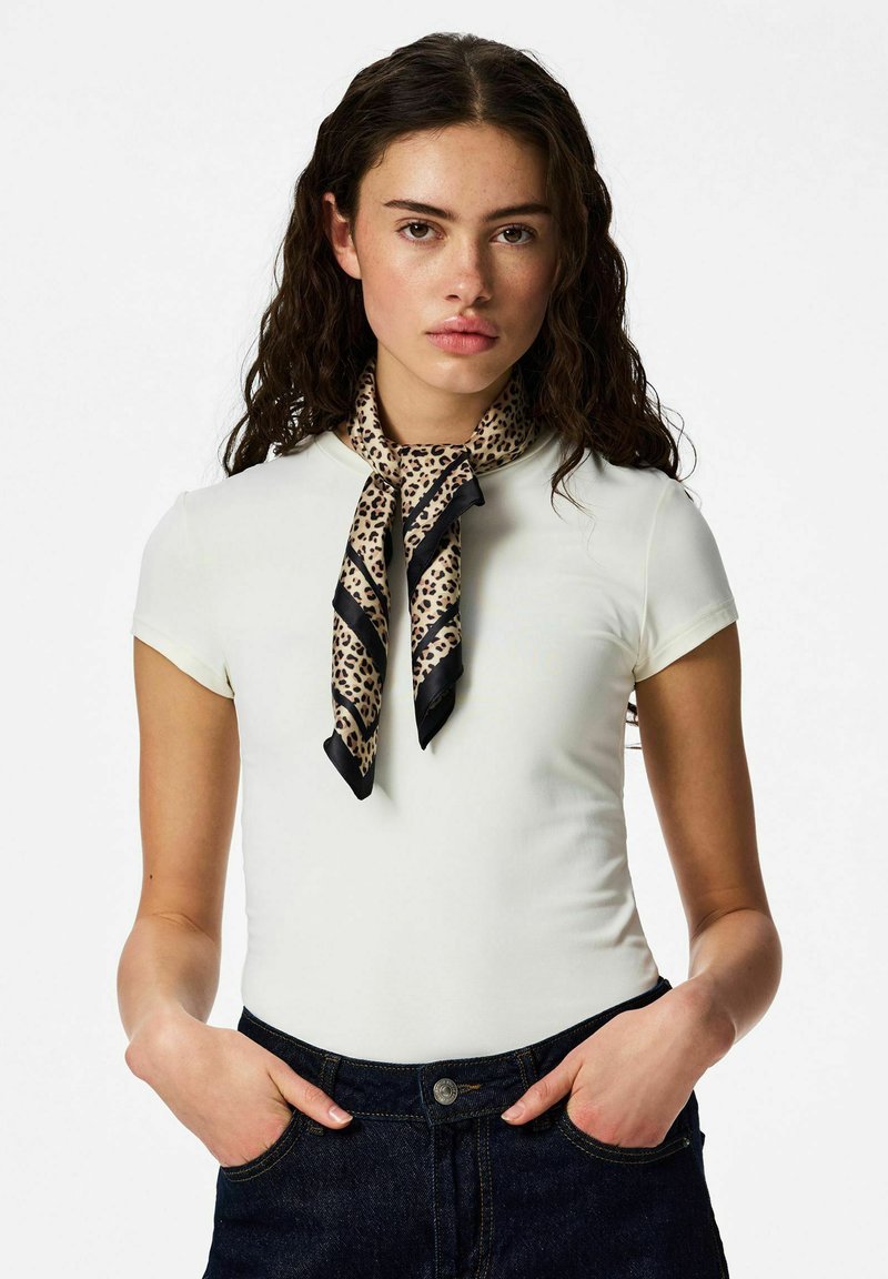 Jeune femme aux longs cheveux bouclés porte un haut blanc à manches courtes, une écharpe à imprimé léopard nouée autour du cou, et un jean bleu foncé avec les mains dans les poches.
