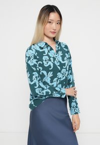 Teal bluse med lyseblå blomsterdesign; langermet, krage og knappet front; kombinert med et mørkeblått skjørt.