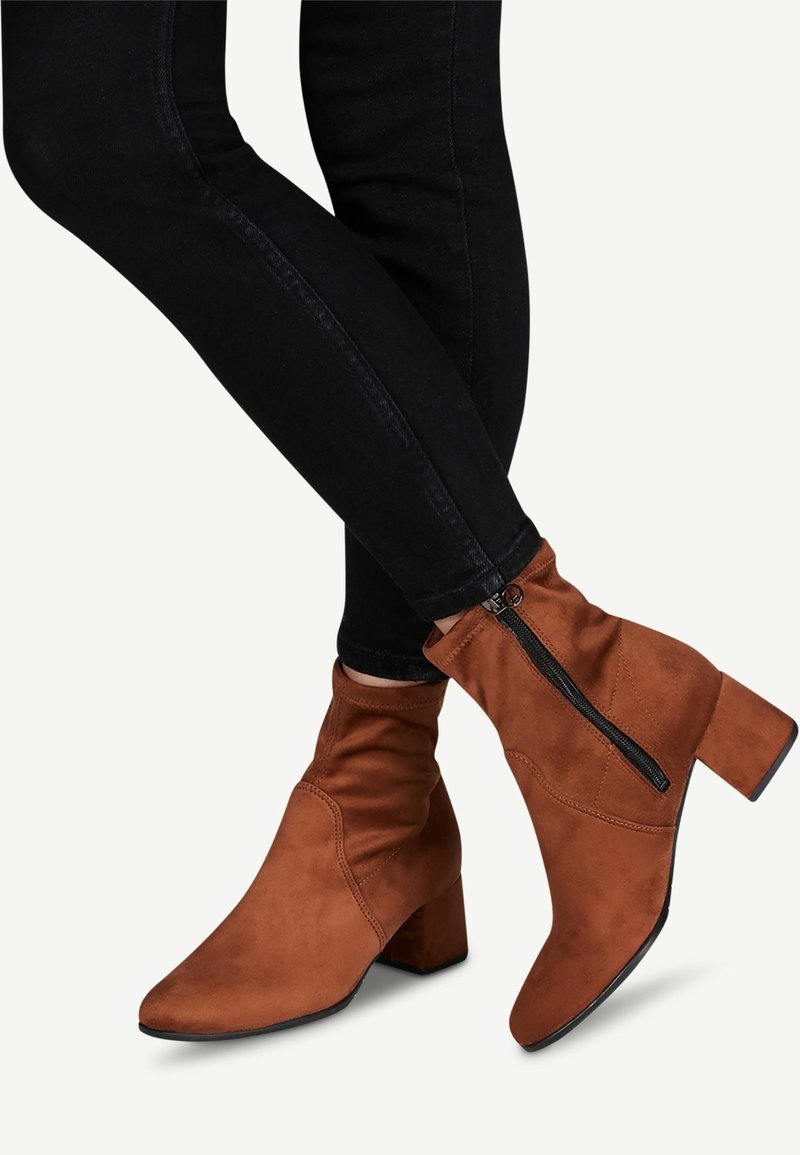 Jambes en pantalon noir moulant portant des bottines en daim marron avec une fermeture éclair sur le côté et des talons blocs, croisées aux chevilles.