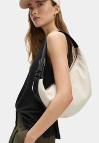 Joven con cabello largo que lleva una gorra beige y una camiseta sin mangas negra, llevando un gran bolso de lona blanca con una correa negra.