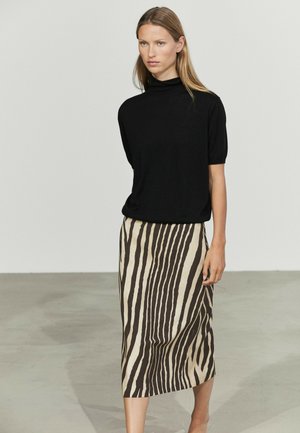 Donna con capelli biondi lisci che indossa un top nero a maniche corte e una gonna midi beige e nera con stampa zebrata, in piedi contro un muro semplice.