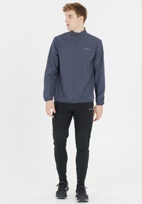 Miestna námornícka vetrovka s vysokým golierom, so zipsom a bočnými vreckami, spojená s čiernymi športovými nohavicami a čiernymi teniskami.