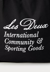 Črna platnena torba z belim besedilom; "Les Deux International Community & Sporting Goods" v različnih stilih in velikostih pisave.
