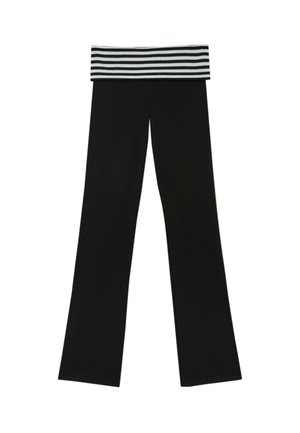 Femme portant un haut à manches longues noir et un pantalon noir avec une ceinture rayée, se tenant avec confiance devant un mur rouge texturé.