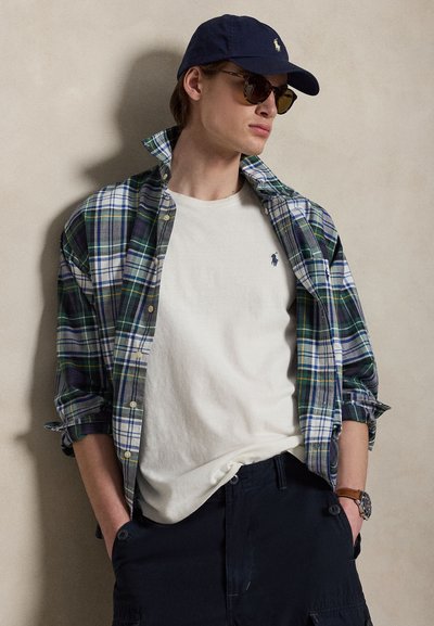 Νέος άνδρας που φοράει μπλε καπέλο, γυαλιά ηλίου, λευκό μπλουζάκι με λογότυπο, πράσινο-μπλε καρό πουκάμισο, σκούρο παντελόνι και ρολόι, στέκεται με τα χέρια στις τσέπες.