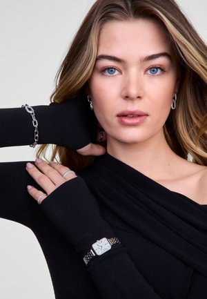 Haut noir à manches longues avec des poignets côtelés, montre argentée avec un cadran rectangulaire et un bracelet en métal, bracelet en chaîne argenté, et plusieurs bagues.