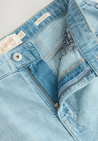 Jean en denim bleu clair avec une coupe décontractée, fermeture éclair et une légère déchirure. L'intérieur présente un accent en tissu à motifs près de la taille.
