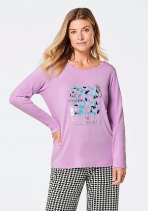 Lila Langarmshirt mit einem grafischen Druck, der blaue und schwarze Akzente zeigt. Kombiniert mit einer Hose im schwarz-weißen Hahnentrittmuster.