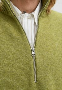 Pogled od blizu na djelomično zakopčanu zeleni pleteni džemper preko bijele košulje s tankim vertikalnim prugama.