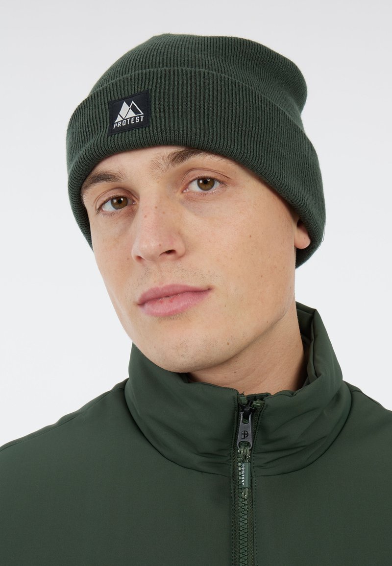 Beanie verde a maglia con texture a costine e risvolto, con un patch nero recante un logo di montagna. Indossato con una giacca verde abbinata.