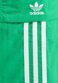 Pantalones cortos verdes hechos de tela ligera con un logo blanco de Adidas y tres rayas verticales blancas a lo largo de los lados.