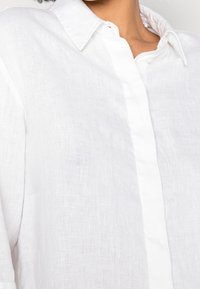 Chemise en lin blanche avec un col boutonné, une texture visible et une patte de boutonnage centrale. Le tissu semble léger et légèrement transparent.