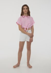 Camicia a maniche corte a righe rosa e bianche legata in vita, abbinata a pantaloni corti bianchi e infradito grigie; sfondo semplice.