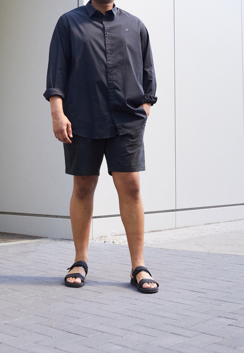 Homme debout en extérieur portant une chemise sombre à boutons, un short noir et des sandales noires sur un trottoir carrelé à côté d'un mur clair.