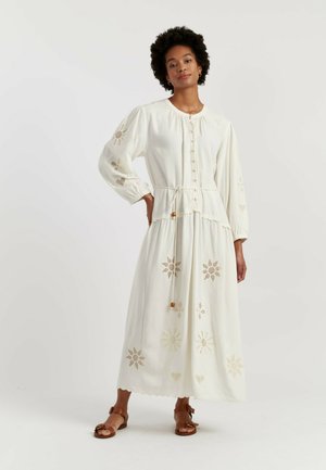 Femme debout, main sur la hanche, portant une longue robe blanche avec un devant boutonné, une taille à cordon et des motifs découpés floraux et en cœur.