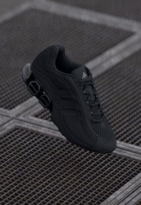 Zwarte sportieve schoen gemaakt van synthetische materialen, met een gestructureerde bovenkant, vetersluiting en drie kenmerkende zijstrepen.