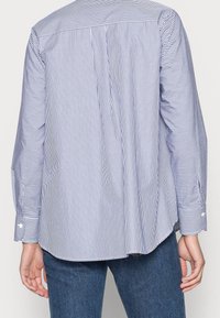 Košile na knoflíky s modrými a bílými vertikálními pruhy, dlouhými rukávy a záhybem na zádech pro lepší tvar. Tkanina působí hladce a lehce.