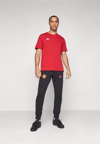 T-shirt vermelho de mangas curtas com o logo branco da Adidas; calças de treino pretas com o logo do Manchester United e detalhes em vermelho nas laterais; sapatilhas de desporto pretas.