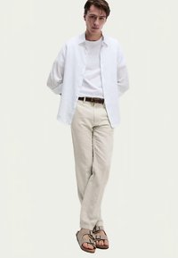 Camisa blanca de lino con botones sobre una camiseta blanca, pantalones beige claro y sandalias grises con tiras. De pie frente a un fondo neutro.