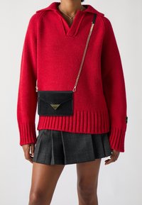 Pull en tricot rouge avec un col polo et un ourlet côtelé, associé à une jupe plissée à carreaux noirs et un petit sac bandoulière noir.