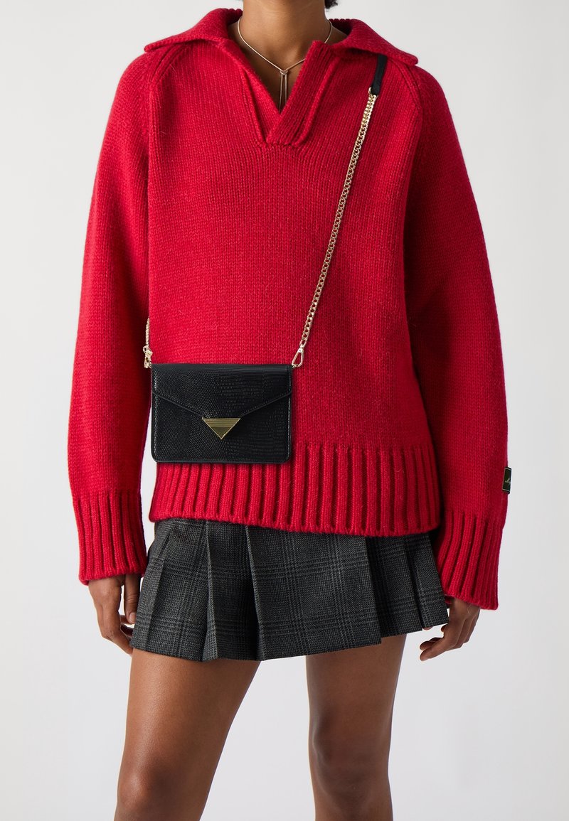 Pull en tricot rouge avec un col polo et un ourlet côtelé, associé à une jupe plissée à carreaux noirs et un petit sac bandoulière noir.