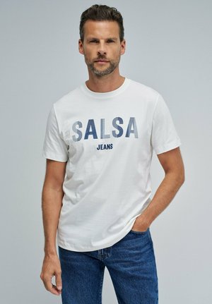 Hombre con camiseta blanca con texto "SALSA JEANS" y vaqueros azules, de pie con una mano en el bolsillo contra un fondo liso.