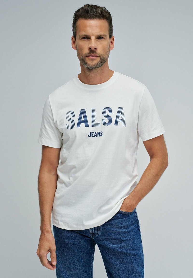 Man draagt een wit T-shirt met de tekst "SALSA JEANS" en een blauwe spijkerbroek, staat met één hand in de zak tegen een effen achtergrond.