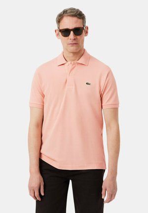 Hombre con gafas de sol negras, camisa polo de manga corta color rosa claro con un pequeño logotipo de cocodrilo verde y pantalones negros.