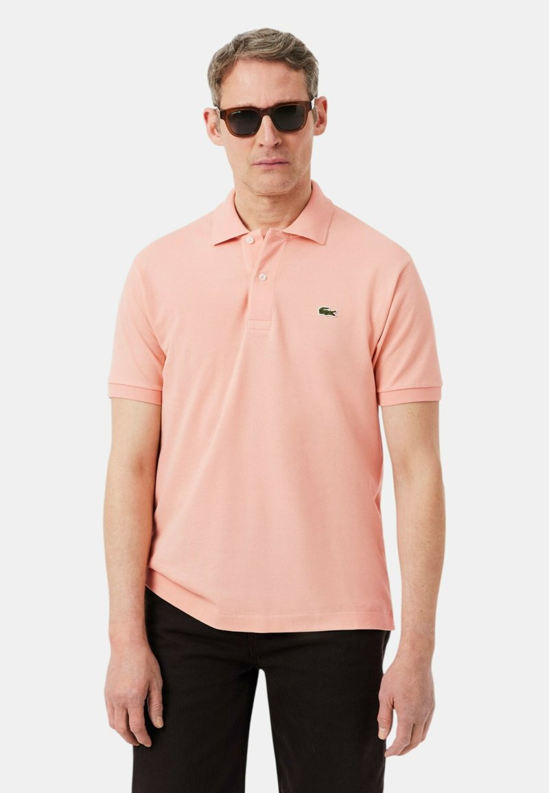 Homme portant des lunettes de soleil noires, un polo à manches courtes rose clair avec un petit logo crocodile vert, et un pantalon noir.