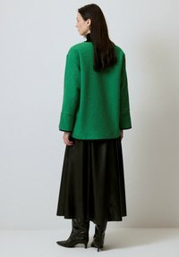 Palton verde texturat cu mâneci lungi, combinat cu o fustă din piele neagră și cizme negre, lucioase, până la genunchi, văzut din spate.