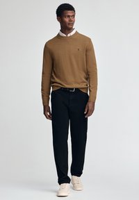 Brązowy sweter dzianinowy z prążkowanym kołnierzykiem, noszony na koszuli z kołnierzykiem, zestawiony z czarnymi spodniami i jasnymi sneakersami. Prosty fason i dopasowany krój.