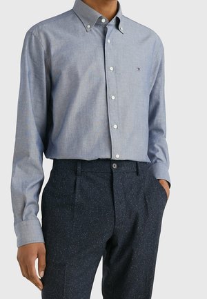 Mann trägt ein hellblaues, geknöpftes Hemd mit kleinem Logo auf der Brust, kombiniert mit dunklen, gesprenkelten Hosen, eine Hand in der Tasche.