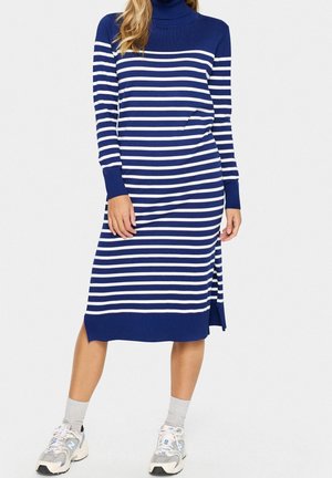 Žena má na sobě pletené šaty střední délky s modrými a bílými vodorovnými pruhy, s dlouhými rukávy a teniskami, na jednobarevném pozadí.