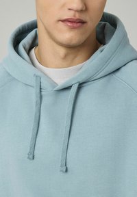 Jeune personne portant un sweat à capuche bleu clair avec des cordons, par-dessus un t-shirt blanc col rond, montrant le bas du visage et le cou.