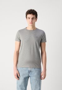 Γκρι, κοντομάνικο μπλουζάκι φτιαγμένο από μαλακό ύφασμα, με στρογγυλή λαιμόκοψη και ένα μικρό λογότυπο στο στήθος. Συνδυασμένο με ανοιχτό μπλε τζιν.