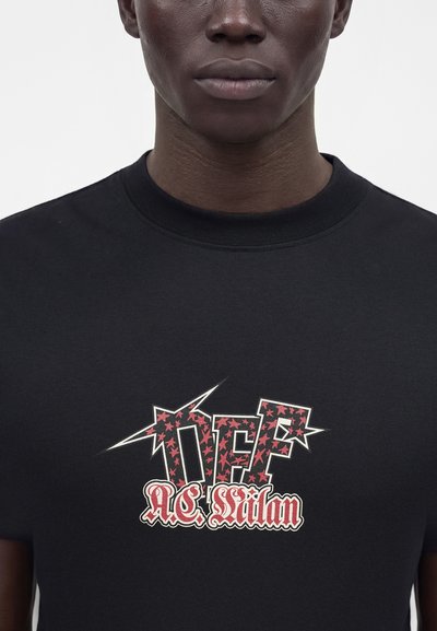 Gros plan d'une personne portant un t-shirt noir avec un logo graphique rouge et blanc indiquant "OFP AC Milan" sur la poitrine.