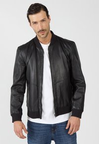 Man in een zwart leren bomberjack over een wit shirt en blauwe jeans, poserend tegen een plain lichte achtergrond.