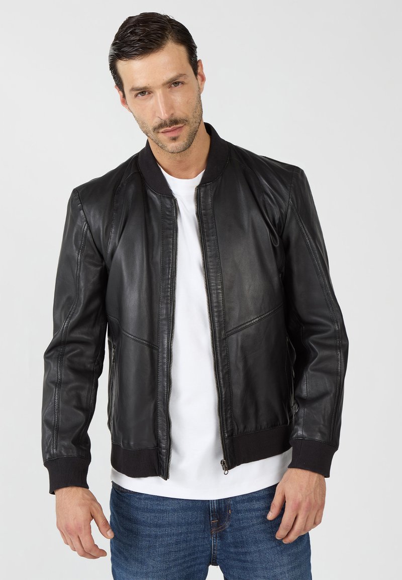 Man in een zwart leren bomberjack over een wit shirt en blauwe jeans, poserend tegen een plain lichte achtergrond.