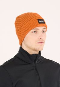 Orange strikket hue med ribbet tekstur og en sort lapp mærket "SLOPE." Båret med en sort zip-neck top.