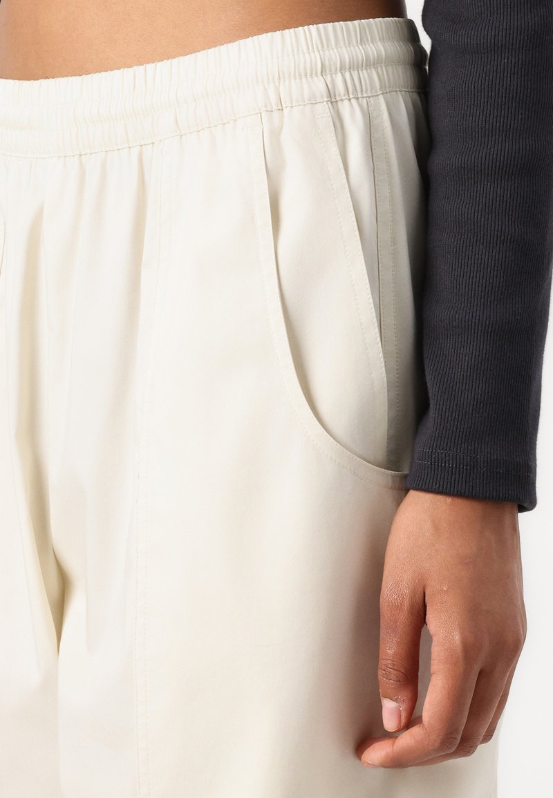 Un pantalon de couleur crème, avec une taille élastique, dispose de poches latérales et d'une texture lisse. Un haut à manches longues de couleur sombre est partiellement visible en haut à gauche.