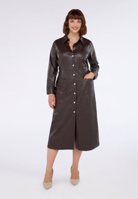 Donna sorridente, che indossa un vestito midi in finta pelle marrone scuro con bottoni e maniche lunghe, abbinato a tacchi a punta beige, in piedi su uno sfondo bianco.