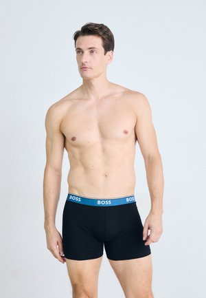Schwarze Boxershorts mit einem blauen "BOSS"-Bund, aus glattem Stoff, enganliegendem Design und mittellanger Länge bis zum Oberschenkel. Modell steht aufrecht.