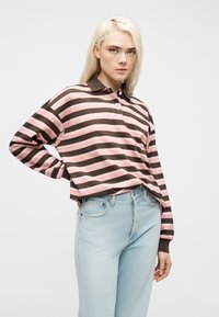 Γυναίκα με μακριά ξανθά μαλλιά, που φοράει ροζ και καφέ ριγέ μπλούζα πόλο με μακριά μανίκια και ανοιχτόχρωμα μπλε τζιν, στέκεται με το ένα χέρι στη μέση.