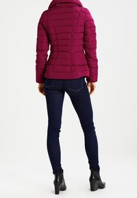 Mujer vestida con jeans ajustados de color azul oscuro, botines negros de tacón y una chaqueta acolchada morada, de pie de espaldas sobre un fondo blanco.