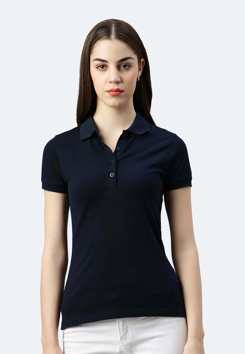 Jeune femme aux cheveux bruns raides portant un polo noir à manches courtes et un pantalon blanc, posant devant un fond clair.